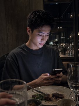 A man in a dark t-shirt looking down at his phone at a restaurant table with a half-eaten plate of food, wine glasses, and a bar in the background.