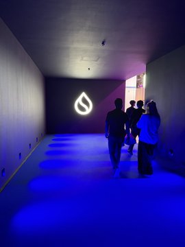 Silhouettes of people walking down a hallway lit with blue and purple floor lights, heading toward an illuminated Sui logo on the wall.