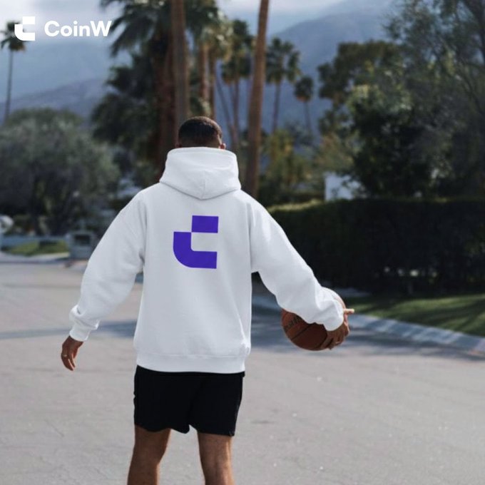 A man viewed from behind walks down a palm-tree-lined street holding a basketball, wearing a white hoodie featuring the purple CoinW logo on the back, with the "CoinW" branding also visible in the top-left corner — tying into the "GM" (good morning) post as a casual, sunlit greeting from the crypto platform.