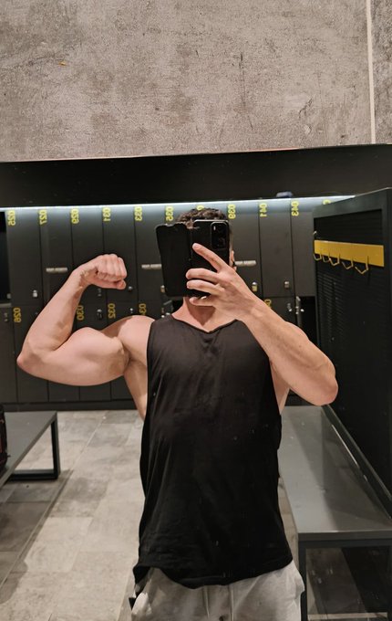 A man in a black tank top taking a mirror selfie in a gym locker room, flexing his right bicep, with numbered lockers labeled "123" through "134" visible behind him.