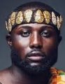 A man with a beard wearing a golden crown-like headpiece adorned with wing-shaped ornaments, with orange flowers visible near his shoulder. The regal crown imagery visually reinforces the post's boastful tone about financial dominance, symbolizing his "king" status and the lavish lifestyle implied by his comments about spending and flexing wealth.
