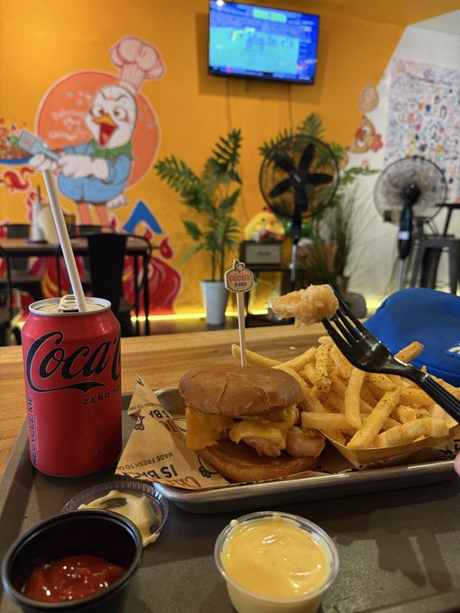A meal of a cheeseburger and crinkle-cut fries served on a metal tray with ketchup and cheese sauce, alongside a can of Coca-Cola Zero, set in a colorful burger restaurant with wall murals.