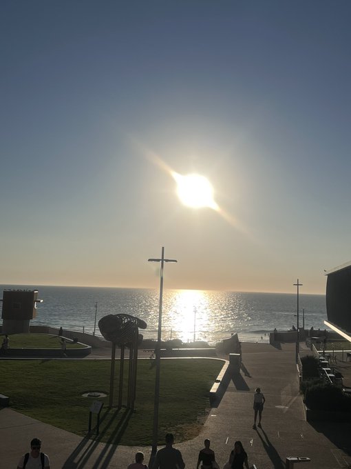 A bright sun shines over a coastal plaza or promenade, casting a reflection on the ocean water while people walk along the path below a cross-shaped monument.