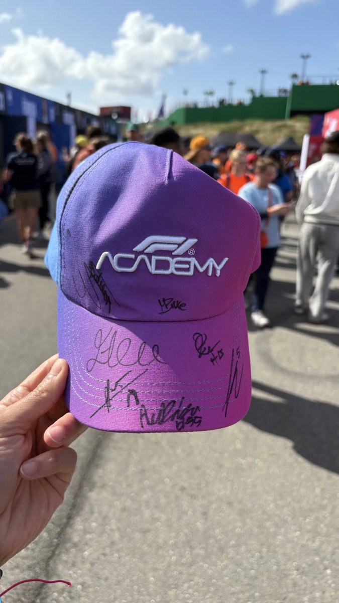 A hand holds up a purple and pink F1 Academy cap with multiple autographs and signatures on it, taken at what appears to be a racing event with crowds in the background.