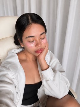 A young woman with dark hair wearing a white hoodie rests her chin on her hand with a tired or pensive expression.