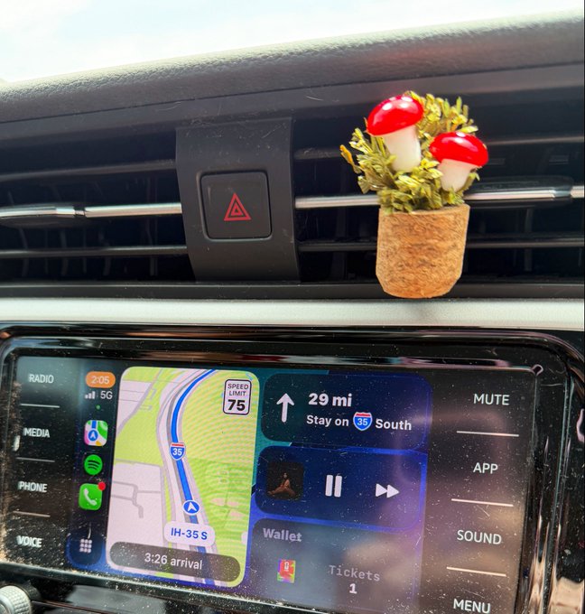 A car dashboard features a GPS display showing directions to stay on I-35 South for 29 miles with a 3:26 arrival time, while a cute decorative mushroom plant sits in the air vent next to the hazard light button. The screen's "Tickets: 1" notification ties into the post's mention of using Avalanche technology to access FIFA World Cup tickets for the Japan vs. Sweden match.