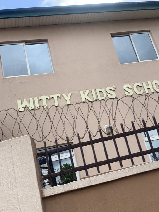 A photograph of a beige building exterior with the gold lettering 'WITTY KIDS SCHO[OL]' partially visible behind a security fence topped with coiled razor wire.