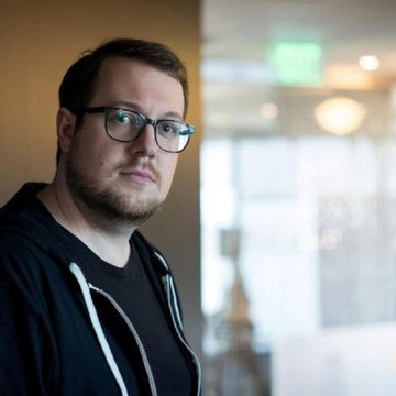 A young man with glasses and a beard, wearing a black hoodie, poses thoughtfully in what appears to be a modern indoor space with blurred glass windows behind him. The image likely depicts Billy Markus, known as Shibetoshi Nakamoto, the co-creator of Dogecoin, whose accidental founder story is being shared in the accompanying post.