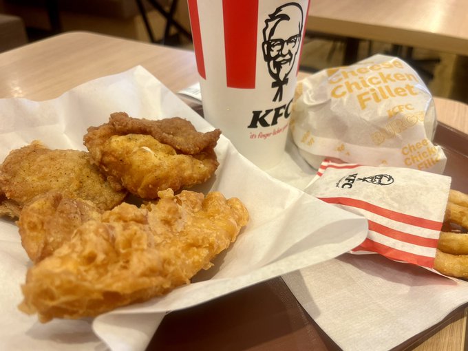 A photo of a KFC meal on a tray, featuring fried chicken pieces in a paper-lined basket, a Cheese Chicken Fillet wrapped sandwich, fries, and a KFC-branded cup with Colonel Sanders' image.