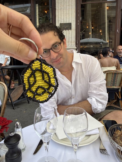 In the image, a man wearing glasses is sitting at a table with wine glasses and a plate of food. A person's hand is holding up a small beaded keychain that features a yellow design with black beads. The caption for this image reads "@xbteneighty pulled up with the coolest handmade @BNBCHAIN keychain from @TheBlockCo has the best team!".