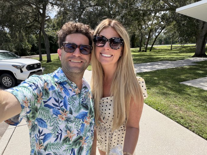 A smiling couple takes a sunny outdoor selfie—the man in a tropical floral shirt and sunglasses, and the woman in a polka-dot dress and dark sunglasses, with a white Toyota SUV parked behind them—celebrating their 9th wedding anniversary while reflecting on a decade of transformation since his release from prison.