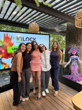 A photograph of five people posing together outdoors under a pergola at an event, standing in front of a screen displaying "UNLOCK SUMMER REWARDS".