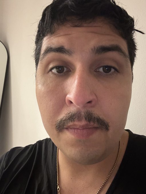 A close-up selfie shows a man with dark curly hair, a mustache, and brown eyes wearing a black v-neck shirt with a thin gold chain necklace. A mirror is partially visible against the plain wall in the background.