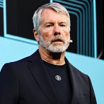 Michael Saylor, the founder of Strategy, is shown speaking at a podium while wearing a dark blazer over a black t-shirt featuring the Strategy "B" logo, with a small microphone attached near his cheek, visually accompanying his statement that "Bitcoin doesn't need any legislation, but the U.S. does."
