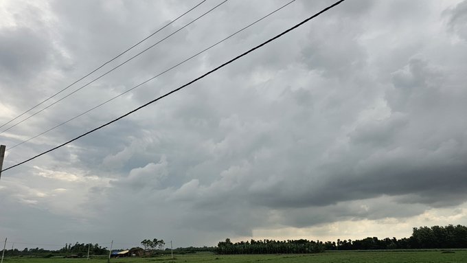 A wide landscape view of dramatic, cloudy skies over a flat green field, with power lines stretching across the upper portion of the frame.