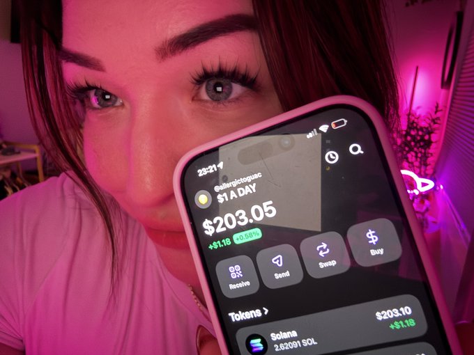 A person with pink/red hair holding up a smartphone showing a crypto wallet app displaying $203.05 (up +$1.18 or +0.56%) with options for Receive, Send, Swap, and Buy, showing a Solana token balance of 2.02001 SOL.