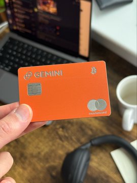 A hand holds up an orange Gemini credit card, with a laptop and coffee mug visible in the background, symbolizing the user's daily Bitcoin purchases funded through crypto rewards earned on the card.