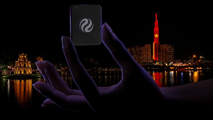 A hand holds up a black square token with a white circular logo against a nighttime city skyline featuring an illuminated red tower with a star and a glowing church-like structure across the water.