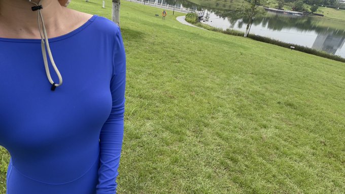 A woman wearing a blue athletic top with a fitness tracker around her neck stands in a grassy field near a pond.