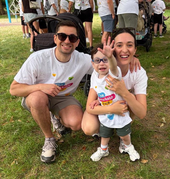 A family of three (two adults and a young child wearing glasses) poses outdoors on grass at what appears to be a 'Remarkable Race' charity event. They are all wearing matching white t-shirts with colorful event logos, and the child is waving at the camera.
