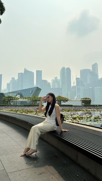 A woman in a white top and pants sits on a wooden bench with a city skyline visible in the hazy background.