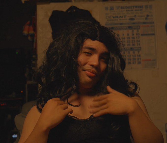 A person with long, dark wavy hair wearing a dark top, posing in a dimly lit room with a calendar visible on the wall behind them.