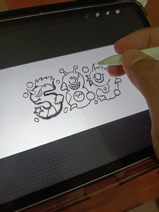 A hand uses a stylus on a tablet screen to draw whimsical cartoon monsters with multiple eyes, horns, and teeth in a sketch app, with a grid pattern visible in the background.