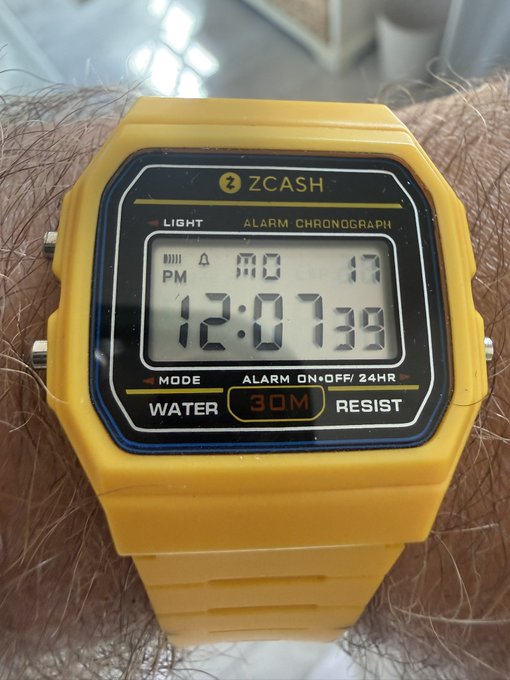 A yellow ZCASH digital wristwatch displaying the time 12:07:39 PM, MO 17, with features labeled including LIGHT, ALARM CHRONOGRAPH, MODE, ALARM ON/OFF/24HR, WATER 30M RESIST.