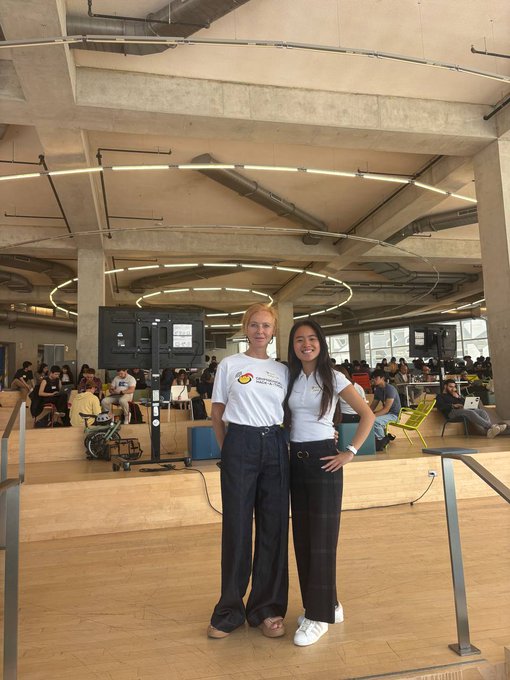 The image shows two women standing next to each other, posing for a picture in front of a large screen TV. They are both wearing white shirts with black lettering that says "LET THEM COOK Workshops kicking off just now at #OpenClaw Hack! Builders about to learn x402, ERC-8004, and GOAT mainnet deployment Fresh AI agents incoming!". The women appear to be enjoying the event or activity taking place in front of them.