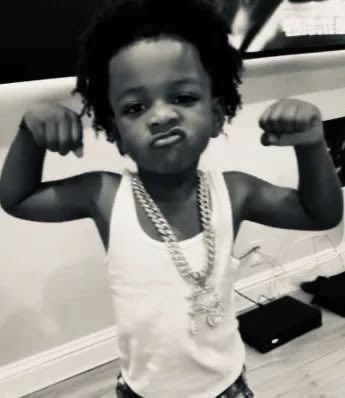 A black-and-white photo of a young child flexing both arms, wearing a white tank top and chain necklace.