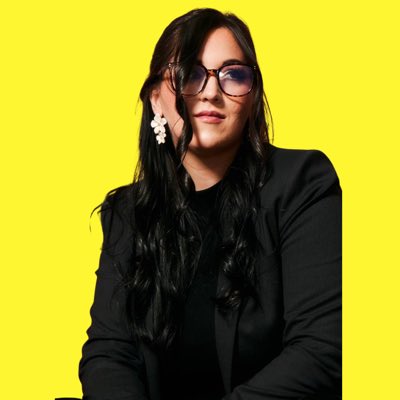 A portrait of a woman with long dark hair wearing black glasses, white flower earrings, and a black blazer against a bright yellow background.