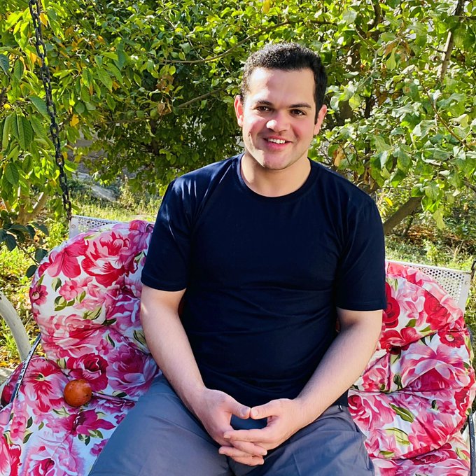 A smiling man in a dark navy t-shirt sits on a floral-patterned cushioned bench in a sunlit garden, surrounded by green foliage.