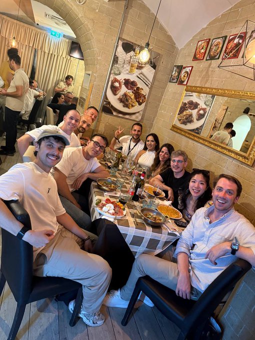 A group of friends share a lively dinner at a restaurant in Naples, posing cheerfully around a table filled with Italian dishes, capturing the spirit of community and collaboration referenced in the post about an emerging partnership between @MonkeDAO and @solana_italy.