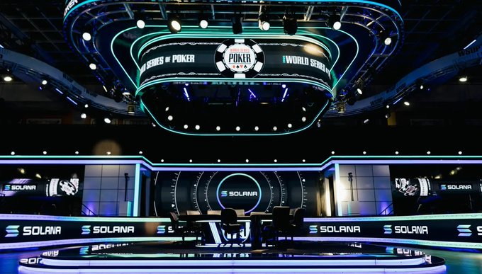 An elaborate, brightly lit poker tournament stage features a large overhead screen displaying 'POKER WORLD SERIES' with 'SOLANA' branding visible throughout the venue.