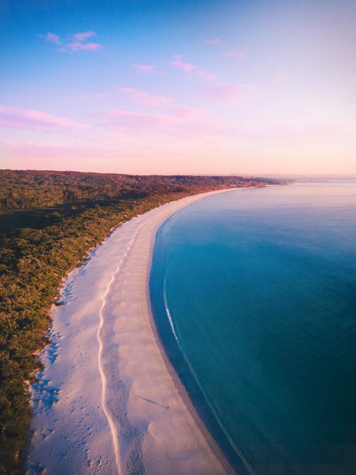 Aerial view of a stunning coastal beach at sunset, featuring a curved white sand shoreline bordered by dense green forest on one side and clear turquoise waters on the other, under a pink and blue pastel sky.
