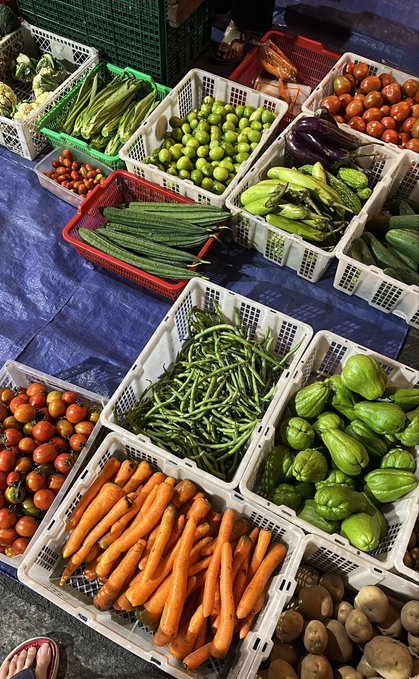 An overhead view of a fresh produce market display with various vegetables arranged in plastic crates, including carrots, long beans, green chayote, cucumbers, tomatoes, eggplants, and broccoli.