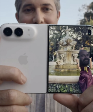 A person holding up a white foldable smartphone displaying a photo of a bearded man in a maroon shirt standing in front of a fountain, with the user's face partially visible above the phone.