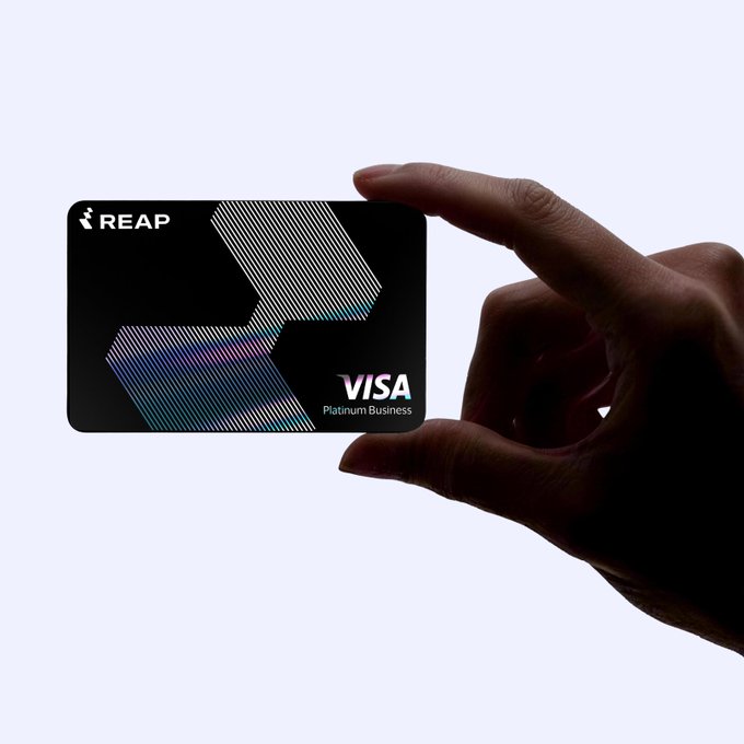 A silhouetted hand holds up a black Reap Visa Platinum Business card, featuring a holographic geometric design. The image illustrates the post about Reap (owned by Kraken's parent company) partnering with Visa to launch stablecoin-funded credit cards across 100+ markets.