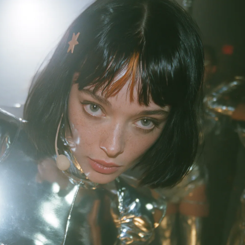 A close-up portrait of a young woman with a dark bob haircut and bangs, wearing a silver metallic outfit and a star hair clip.