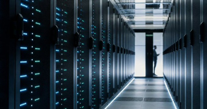 A dark server room with rows of tall server racks illuminated by blue lights, with a silhouette of a person standing in the brightly lit doorway at the end of the aisle.
