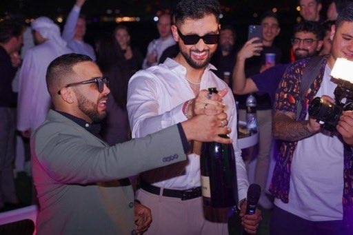A man in a green suit jacket and sunglasses celebrating at a party, opening a large bottle of champagne while surrounded by people in the background, one holding a camera with a flash.