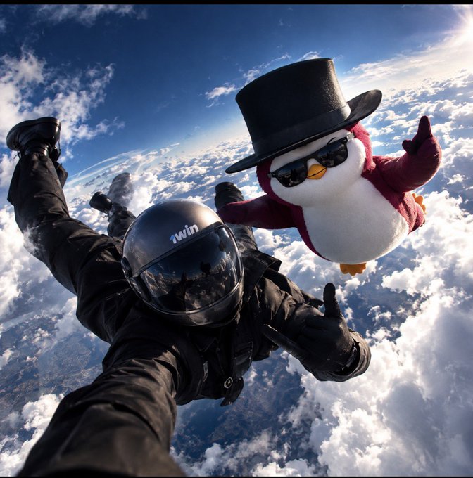 A skydiver in a black jumpsuit and helmet with 'iwin' written on the visor free-falls through the clouds alongside a plush penguin toy wearing a top hat and sunglasses.