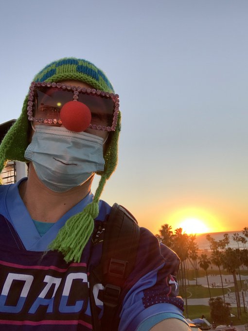 A selfie of a person wearing a clown disguise with a green knit hat, large pink jeweled glasses, red nose, and face mask, taken at sunset with palm trees visible in the background.