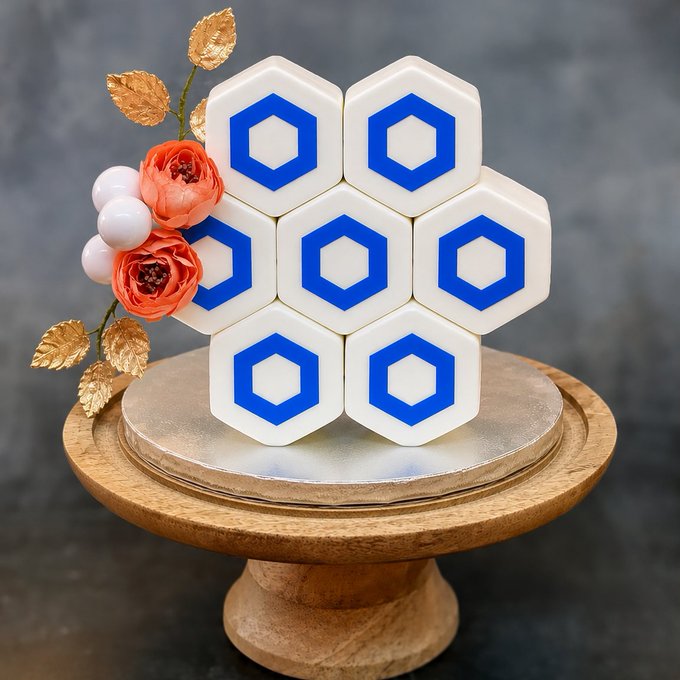 The image features a cake with blue hexagonal shapes, possibly made of fondant or another type of edible material. The cake is placed on a wooden cake stand against a black background. There are also some flowers visible near the bottom left corner of the image. The text "hbd chainlink" appears in the image as well.