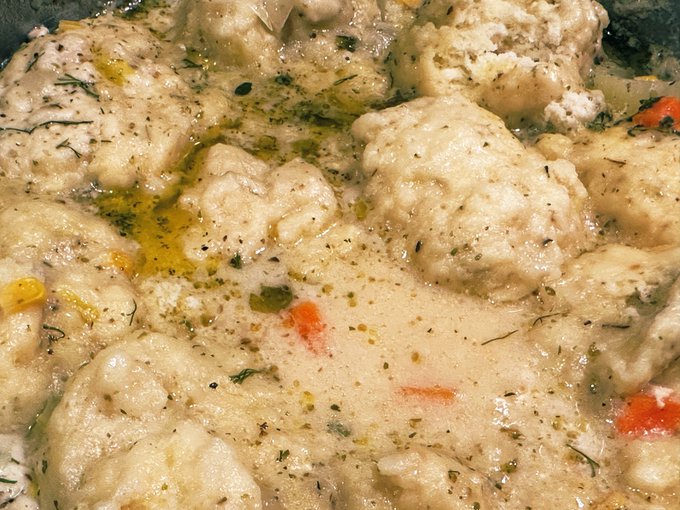 A close-up overhead shot shows a creamy chicken dish, likely chicken and dumplings, garnished with herbs and small pieces of carrots in a pan.