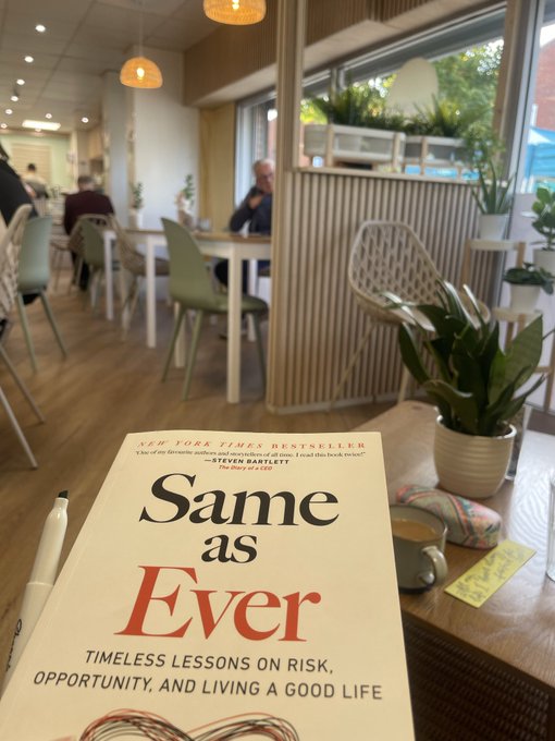 A book titled 'Same as Ever: Timeless Lessons on Risk, Opportunity, and Living a Good Life' rests on a table in a cafe setting with chairs, plants, and a coffee mug visible.