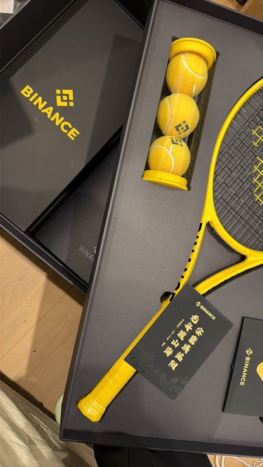 The image features a tennis racket, three yellow tennis balls, and a black box with the words "Binance" written on it. The tennis racket appears to be made of carbon fiber and has a black handle. There are also two small white cards placed next to the tennis racket in the box. The post mentions that even remote areas in Australia can receive these gift boxes as part of Binance's Dragon Boat Festival, which is an event held annually in China to celebrate the traditional race involving rowboats and oars.