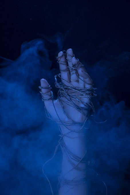 A dark, atmospheric image of a pale hand wrapped in wire, rising through swirling blue smoke against a black background.