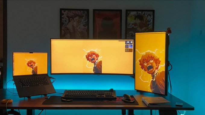 A multi-monitor gaming setup displays an anime character across a laptop and two external screens, with anime posters on the wall and teal LED backlighting illuminating the desk.