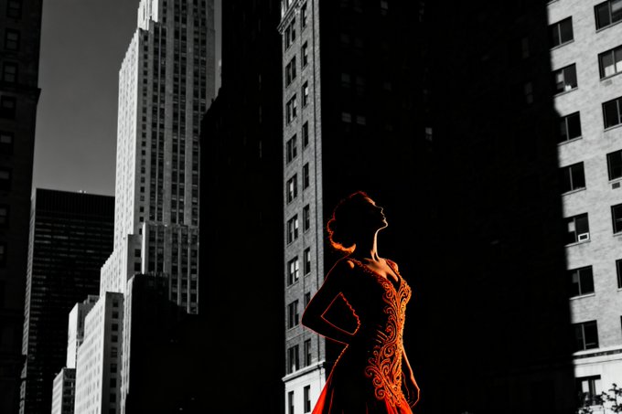 A woman in a red dress stands silhouetted against tall city buildings, lit by the warm glow of the sun.